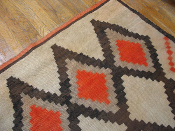 Beige flatweave rug with orange and brown stepped patterns lying diagonally on light wood flooring.