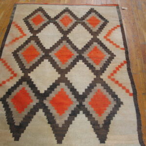 Cream flatweave rug with a geometric stepped pattern of orange and dark brown diamonds on wood plank flooring.