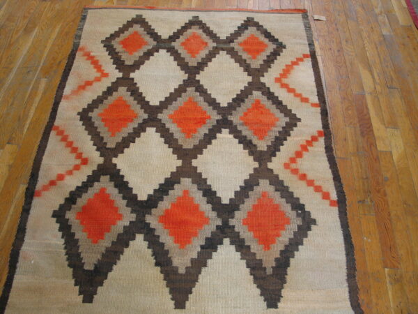 Cream flatweave rug with a geometric stepped pattern of orange and dark brown diamonds on wood plank flooring.