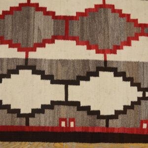 A flatweave rug with stepped diamond patterns in cream, gray, dark brown, and red rests on a wood floor.