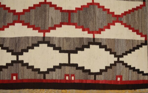 A flatweave rug with stepped diamond patterns in cream, gray, dark brown, and red rests on a wood floor.