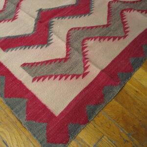 Corner view of a flatweave rug with cream, gray, and pink sawtooth zigzag patterns over wood.