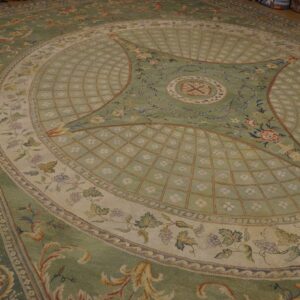 A sage green, cream, and gold low-pile rug with elaborate classical floral patterns rests on a wood floor.