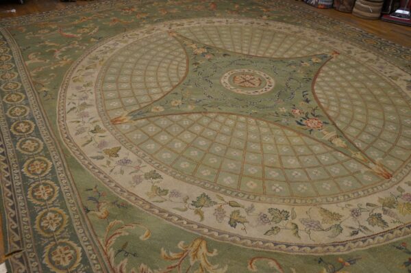 A sage green, cream, and gold low-pile rug with elaborate classical floral patterns rests on a wood floor.