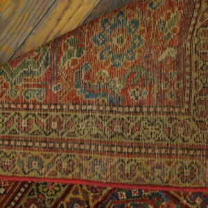 A richly patterned red and blue rug, partially rolled, lies on distressed yellow wood planks.