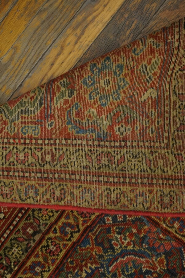 A richly patterned red and blue rug, partially rolled, lies on distressed yellow wood planks.