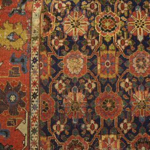Close-up of a low-pile rug with navy, red, green, and gold floral patterns resting on light wood planks.