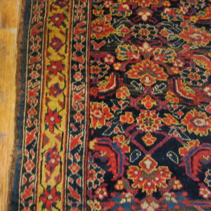 High pile rug with red, yellow, and navy traditional motifs and border lying next to light wood flooring.