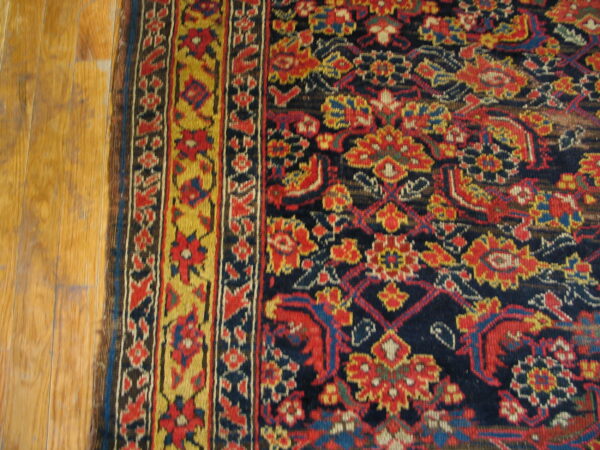 High pile rug with red, yellow, and navy traditional motifs and border lying next to light wood flooring.