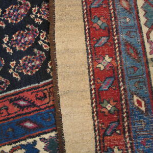 Dark patterned rug pile with red and blue borders folded to reveal the light tan woven backing.