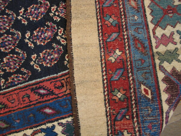 Dark patterned rug pile with red and blue borders folded to reveal the light tan woven backing.