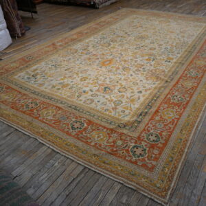 Cream and rust-colored traditional floral rug displayed on aged wooden plank flooring in a warehouse.
