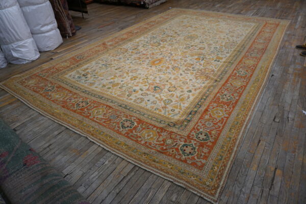 Cream and rust-colored traditional floral rug displayed on aged wooden plank flooring in a warehouse.