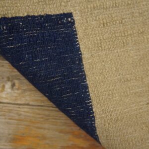 A flatwoven tan rug with a navy blue stripe is partially curled over rustic wood flooring.