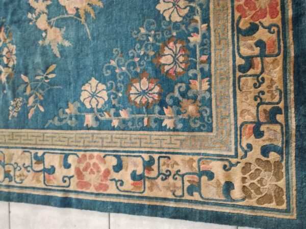 Teal rug with tan and pink floral motifs, featuring multi-layered borders on a visible tiled floor.