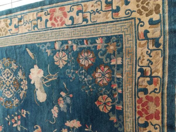 Teal rug corner featuring intricate tan, pink, and blue floral motifs with a detailed greek key border.
