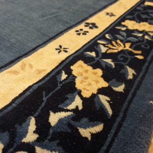 A navy and pale yellow floral border edges a denim blue high-pile rug lying on wood plank flooring.