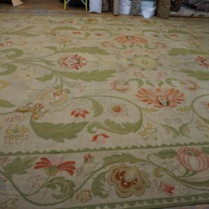 Beige rug with large green leaves and colorful florals laid on a wood floor near stacked rolls.