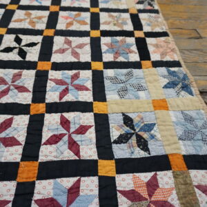 Quilted patchwork textile with multi-colored pinwheel stars framed by black and orange strips on rustic wood planks.