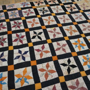 Patchwork textile featuring colorful pinwheel star blocks framed by dark and orange borders on distressed wood flooring.