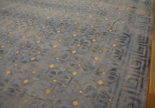 Muted blue and gray rug with gold diamonds, floral motifs, and a greek key border on wood floor.