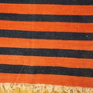 Orange and dark striped flatweave rug with white fringe resting on a light wood floor.