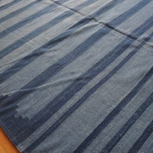 Blue flatweave rug features wide vertical stripes of dark and light denim blue on hardwood floor.