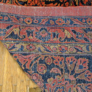 A low-pile traditional rug corner, featuring blue and terracotta floral patterns, is lifted over hardwood flooring.