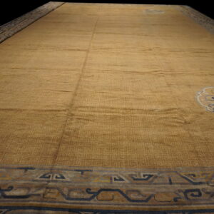 Low-angle view of a gold rug with tone-on-tone fretwork, stylized medallions, and navy blue borders.