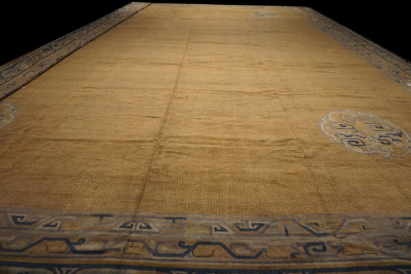 Low-angle view of a gold rug with tone-on-tone fretwork, stylized medallions, and navy blue borders.