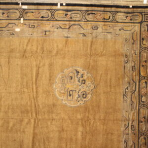 Wrinkled ochre rug with detailed black borders and a central motif rests on light wood floors.