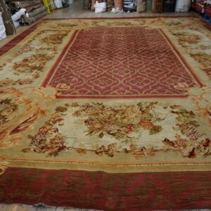 Traditional red and tan rug with floral borders displayed on a wood floor near many rolled textiles.