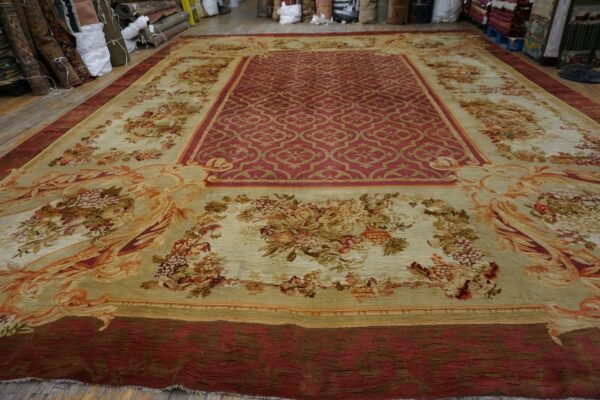 Traditional red and tan rug with floral borders displayed on a wood floor near many rolled textiles.
