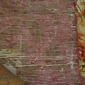 The beige and orange pile rug is folded, exposing the striped maroon and brown backing on wood planks.