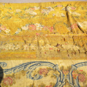Damaged yellow floral rug fragment on a gray surface, showing pink and blue patterns and a large dark shadow.