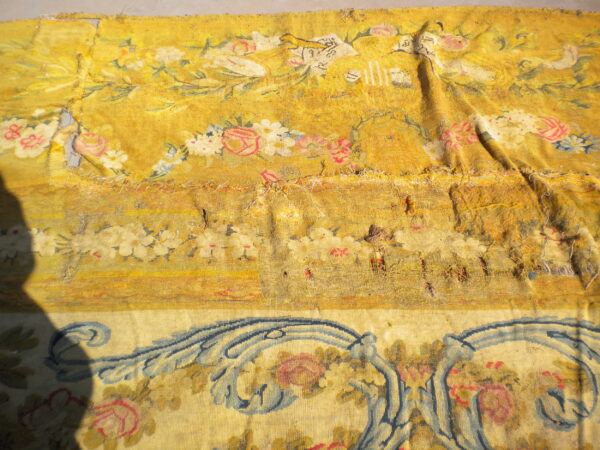 Damaged yellow floral rug fragment on a gray surface, showing pink and blue patterns and a large dark shadow.