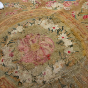 Close view of a worn floral rug in tan and pink with extensive pile loss; white shoes are visible.