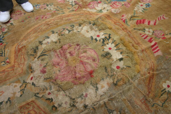 Close view of a worn floral rug in tan and pink with extensive pile loss; white shoes are visible.