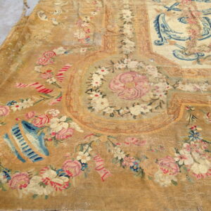 Heavily worn tan low-pile rug showing pink roses, blue scrolls, and floral garlands on a cement floor.