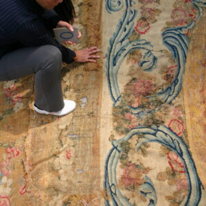 Woman examines a heavily damaged low-pile rug featuring faded floral motifs and dark blue scrollwork.
