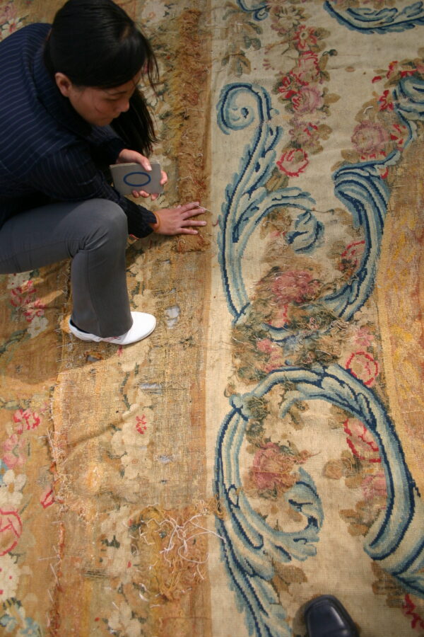 Woman examines a heavily damaged low-pile rug featuring faded floral motifs and dark blue scrollwork.