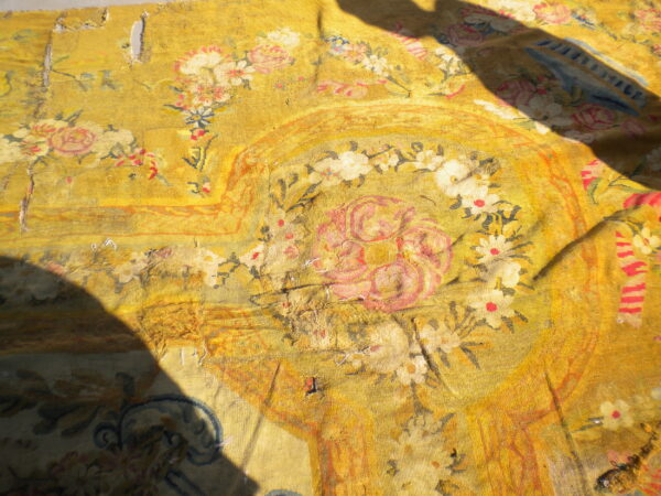 Worn yellow textile fragment with pink and white floral designs, visible damage, and large dark shadows.