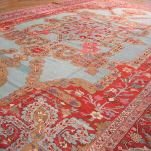 Ornate traditional rug featuring a light blue field and complex red and tan borders on a wood floor.