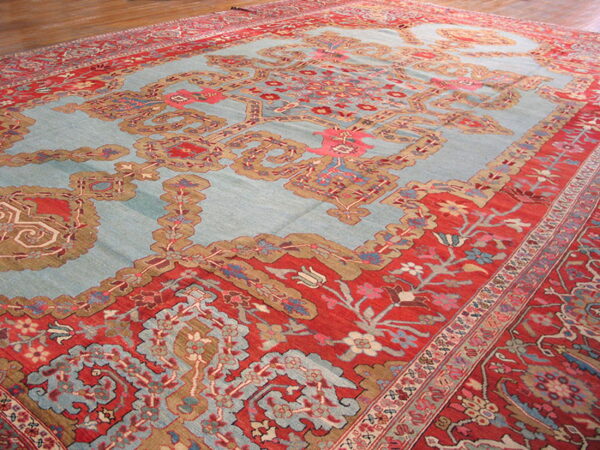 Ornate traditional rug featuring a light blue field and complex red and tan borders on a wood floor.