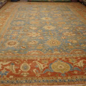 Ornate light blue and rust red floral rug on a wooden floor with rolled rugs visible in the background.