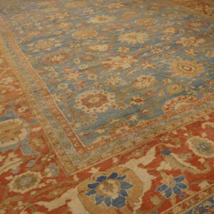 Low-pile traditional rug featuring a faded blue floral field and a wide terracotta red border on wood flooring.