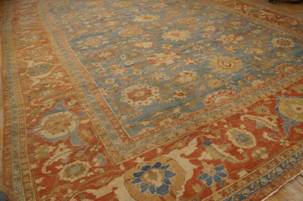 Low-pile traditional rug featuring a faded blue floral field and a wide terracotta red border on wood flooring.