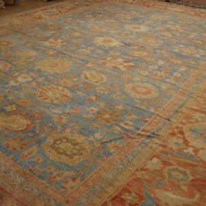 Traditional blue, gold, and rust floral rug displayed on hardwood floors alongside other stacked rugs.