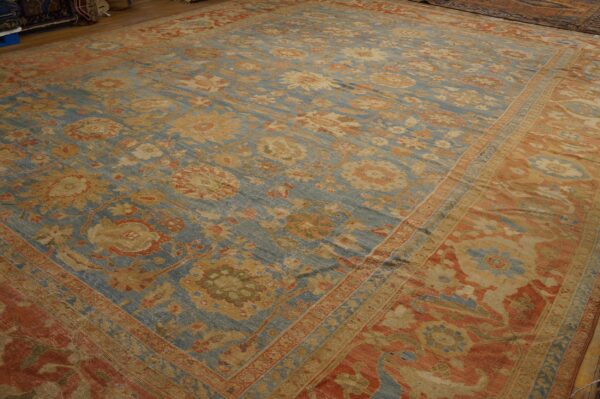 Traditional blue, gold, and rust floral rug displayed on hardwood floors alongside other stacked rugs.