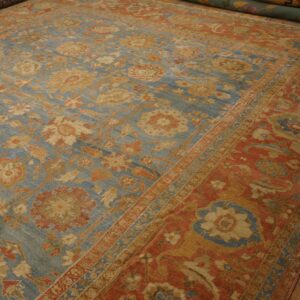 Muted blue floral rug with rust accents displayed on hardwood flooring next to rolled rugs.